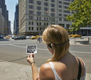 Image of a participant in a Janet Cardiff walk holding a photo of a city at an earlier time, up against the present day city, wearing headphones, and walking forward.